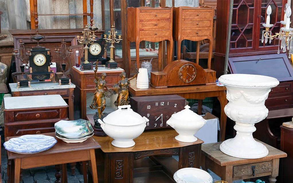 A collection of fine and antique wooden furniture and porcelain pots and dishware sit in a yard sale on a sunny day