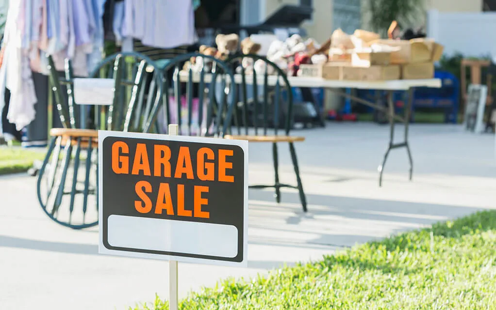 A sign with the words "GARAGE SALE" sits in a home's front yard as belongings that are for sale sit in the driveway of a home in the background on a sunny day