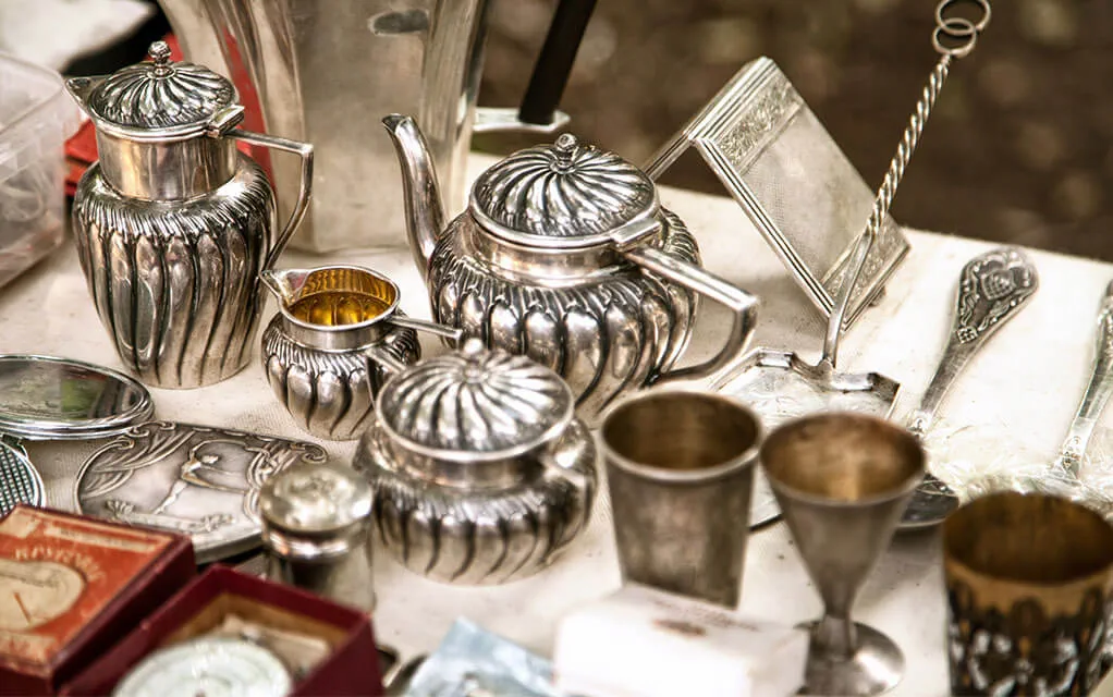 A set of fine, silver colored tea pots and other miscellaneous items sits on a table in a garage sale ready to be bought
