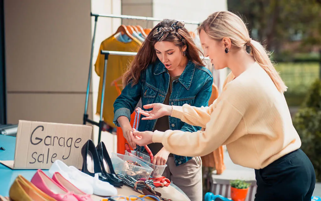 A couple of young women peruse a garage sale set up, looking through some shoes on a table