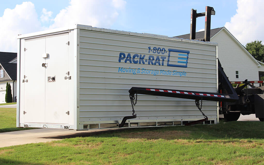 Side view of a 1-800-PACK-RAT container being placed in the driveway of a neighborhood home by the mini-mover lift system on a bright and sunny day