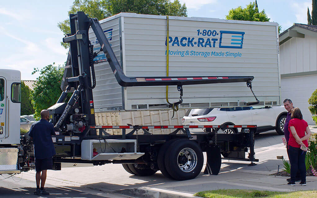 A 1-800-PACK-RAT container is being delivered to a customer's home as the customer's watch a 1-800-PACK-RAT mini-mover driver move the container off the truck and into the driveway