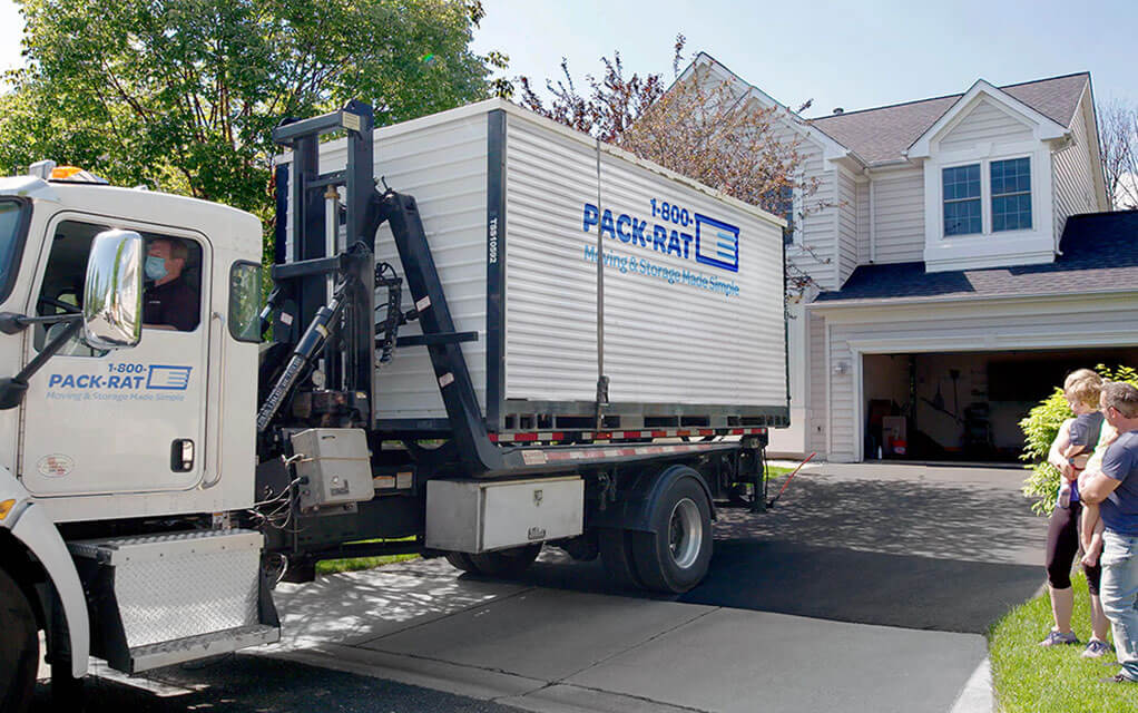 A 1-800-PACK-RAT mini-mover truck is backing into the driveway of a customer's home as they watch on a sunny day
