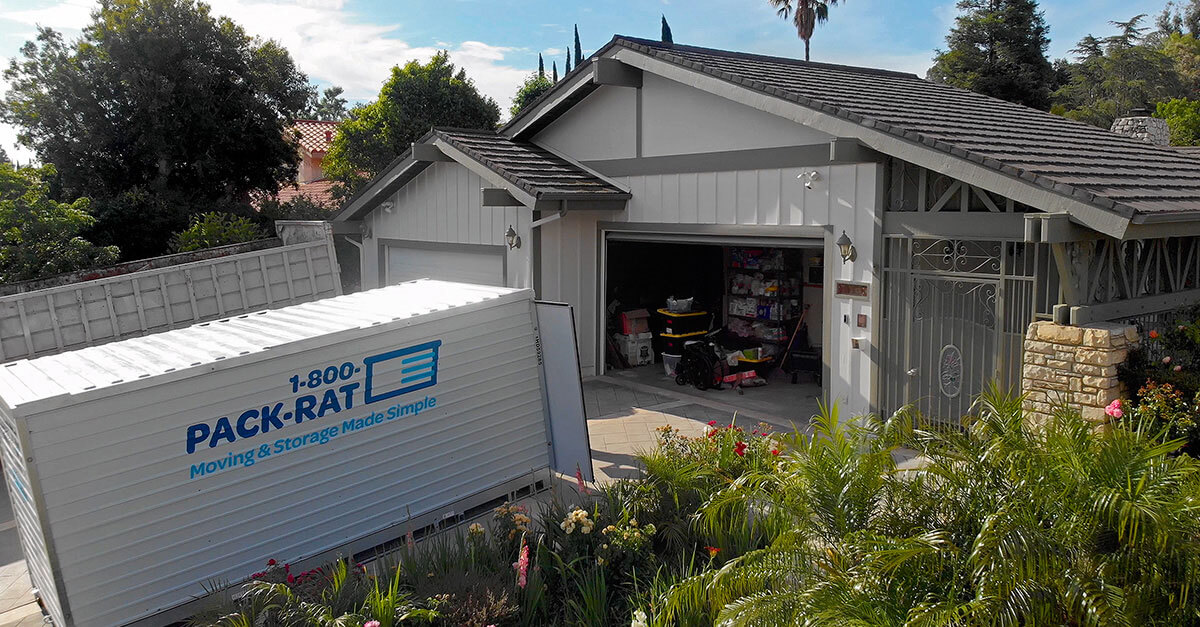 A 1-800-PACK-RAT sits in the driveway of a home with its garage open as the container is open and facing the home being packed by the owners