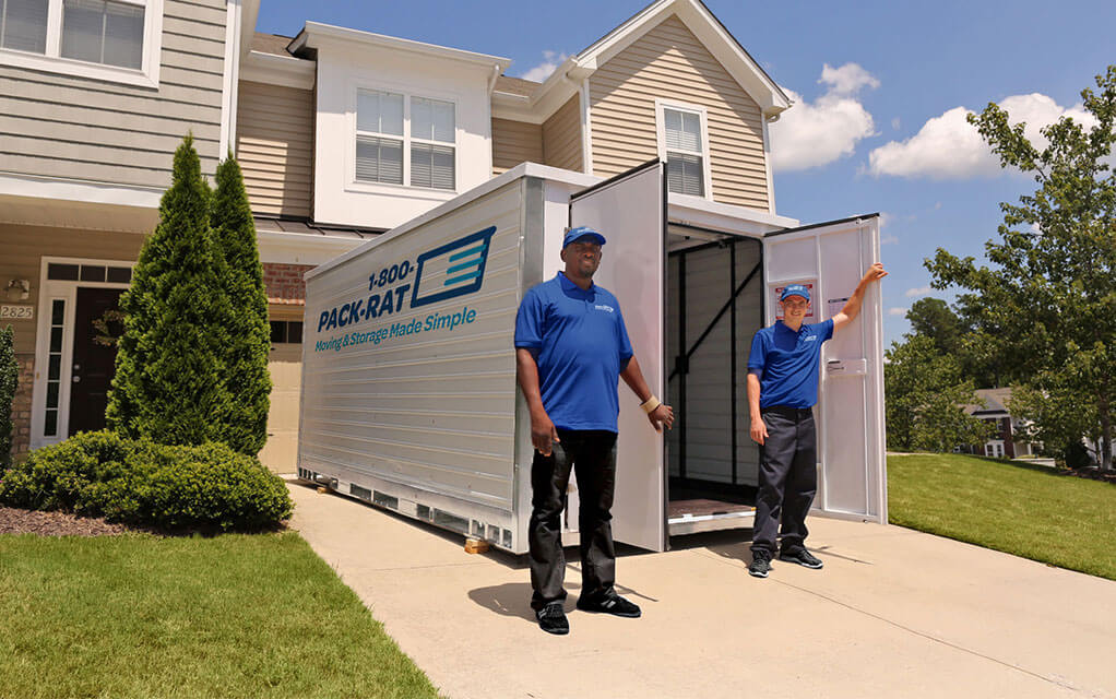 Two men wearing 1-800-PACK-RAT shirts stand in front of an open 1-800-PACK-RAT portable storage container in the driveway of a home on a bright and sunny day