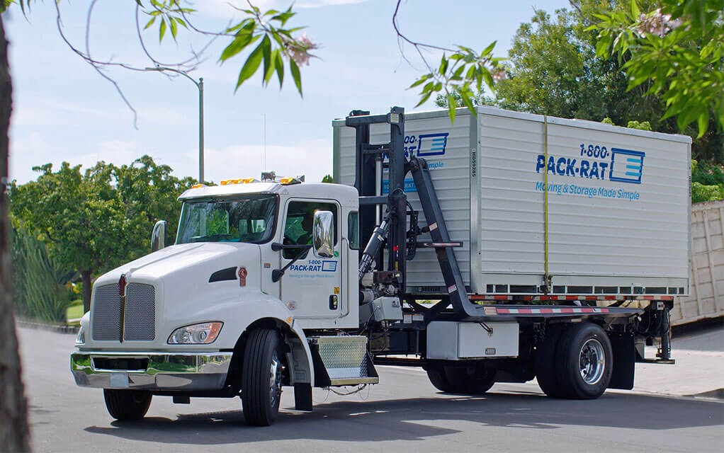 A 1-800-PACK-RAT mini-mover truck is driving a customer's container to their location on a bright and sunny day