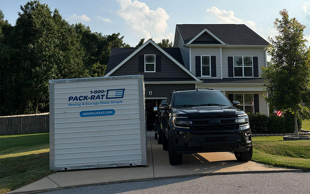 The back view of a 1-800-PACK-RAT portable storage container sitting in the driveway of a suburban home next to a large SUV on a sunny evening