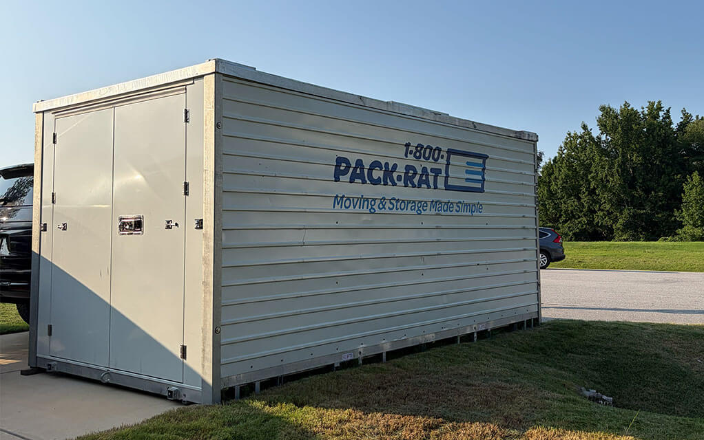 The side view of a closed 1-800-PACK-RAT portable storage container sitting in the driveway of a suburban home on a sunny evening