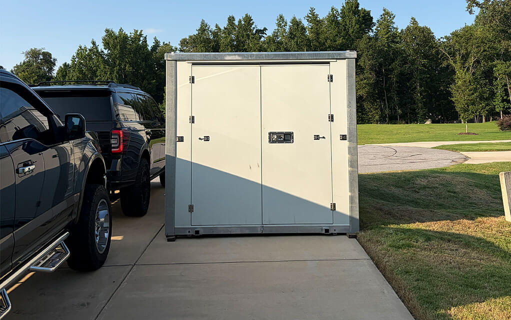The front view of a closed 1-800-PACK-RAT portable storage container sitting in the driveway of a suburban home next to a large SUV on a sunny evening