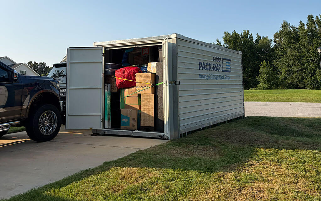 The front corner view of an open 1-800-PACK-RAT portable storage container sitting in the driveway of a suburban home next to a large SUV on a sunny evening with home belongings and boxes packed securely inside
