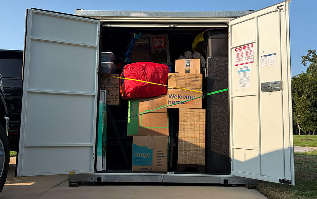 The front view of an open 1-800-PACK-RAT portable storage container sitting in the driveway of a suburban home next to a large SUV on a sunny evening with home belongings and boxes packed securely inside