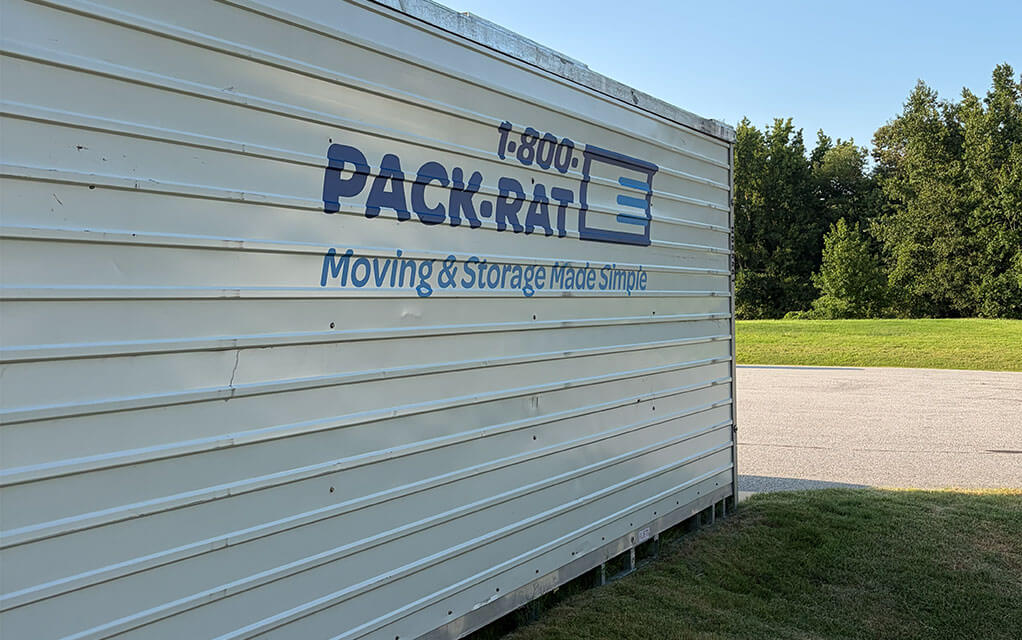 Side view of a 1-800-PACK-RAT portable storage container with the logo clearly visible as it sits in the driveway of a suburban home on a bright and sunny evening