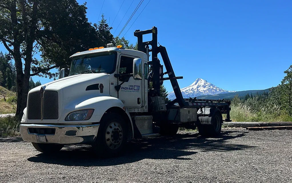 A 1-800-PACK-RAT mini-mover truck with no container on it sits in a parking lot on a clear and sunny day as a mountain top covered in snow is in the background
